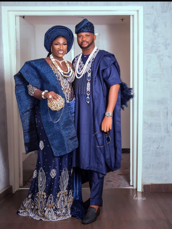 Couple In Traditional Nigerian Blue Lace Attire Posing Together Against A Neutral Background.